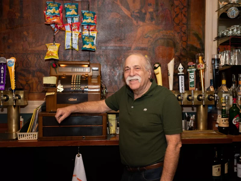 A bald man with a large silver mustache stands at an antique cash register near tap handles and bottles of liquor