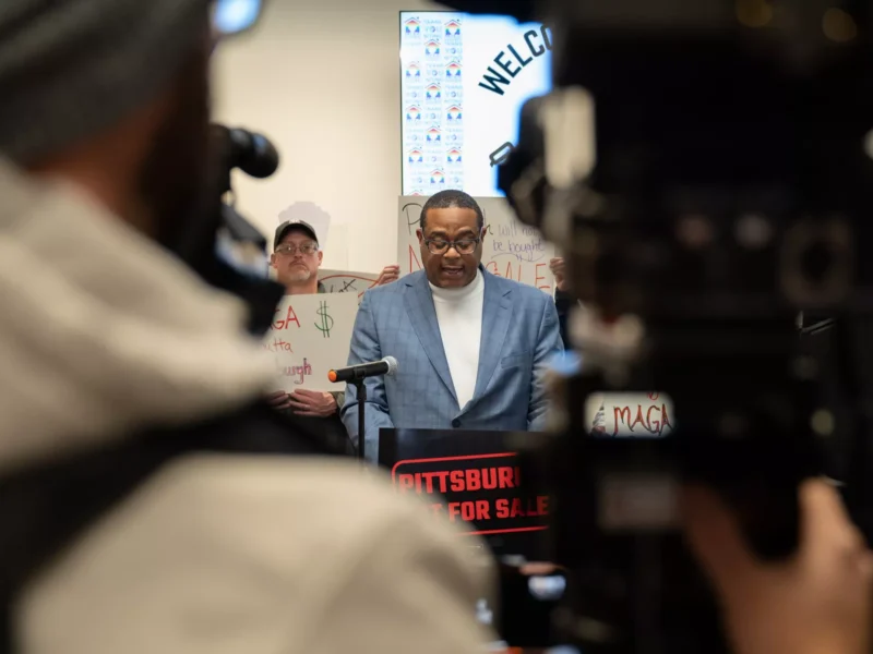 Ed Gainey wears glasses, a turtleneck, and a plaid suit jacket and stands behind a podium reading "Pittsburgh is not for sale" in a crowded room
