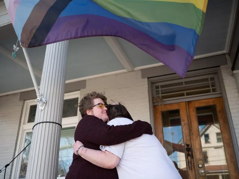A person in a sweater with sideburns, glasses, and a lip piercing hugs a younger person on a porch under a Pride flag