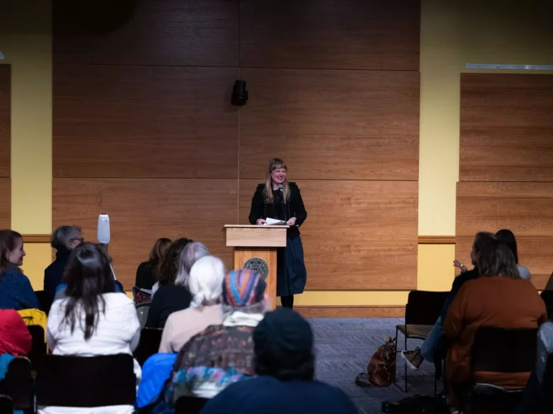 A woman in a black dress with long blond hair smiles as she addresses an auditorium full of women writers