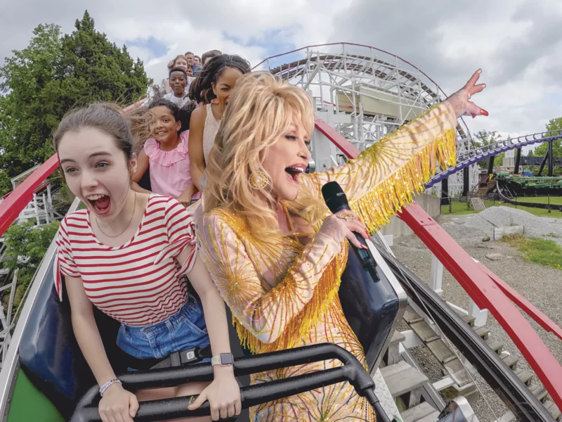 A picture of Dolly Parton superimposed onto Kennywood's rollercoaster, The Thunderbolt next to a little girl in a striped shirt.