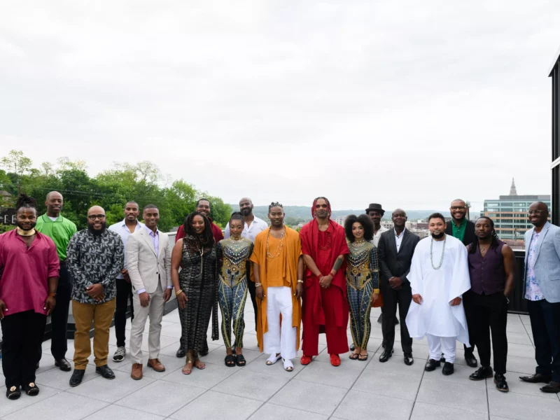 A group of Black artists in a variety of traditional and richly patterned clothes stand on a rooftop terrace