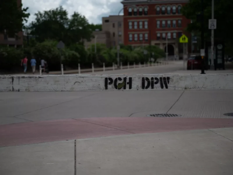 A gentle red-tinted slope of concrete sticks out from a rounded curb behind a wooden Department of Public Works barrier
