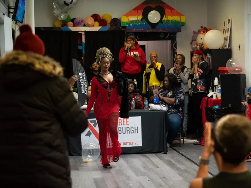 A dancer in drag struts her stuff at the QMNTY Center in front of a sign that says, "World Aids Day."