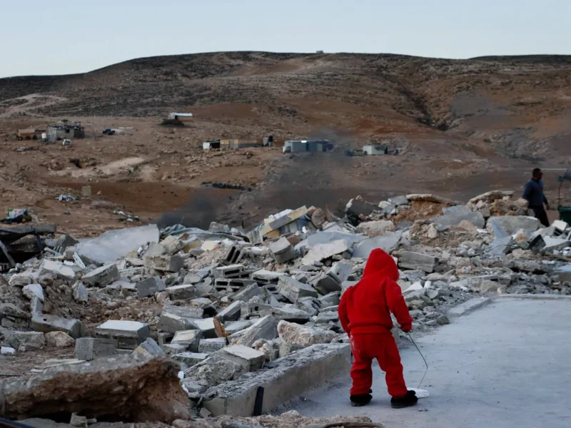 A child is seen from behind standing amid rubble and poking an object with a stick in a hoodie, pants and slippers