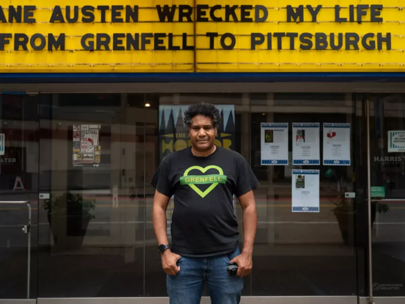 A man wearing a shirt bearing GRENFELL in a green heart stands under a movie theater marquee