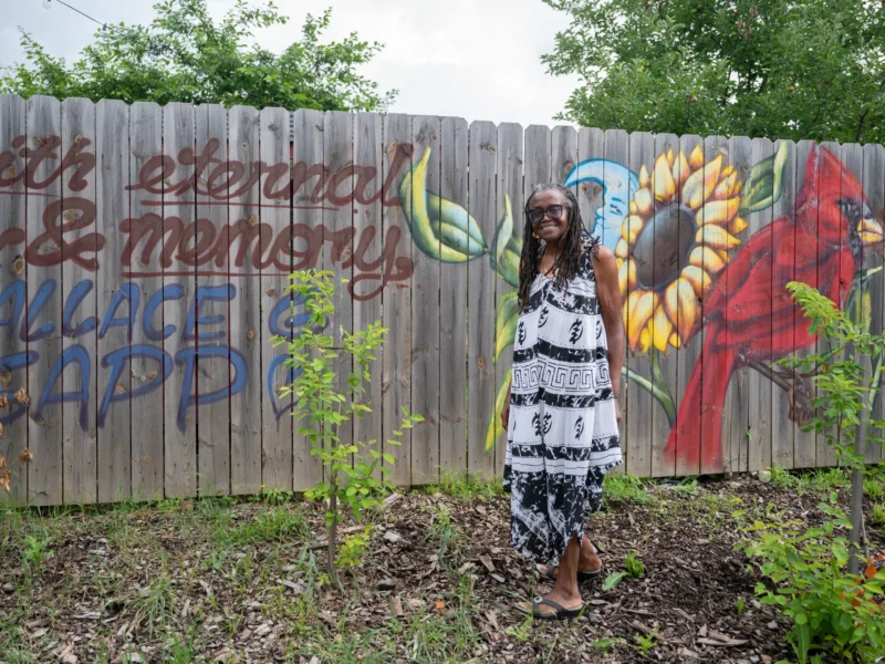 A tall woman with dreadlocks and wearing a flowing dress stands among growing crops