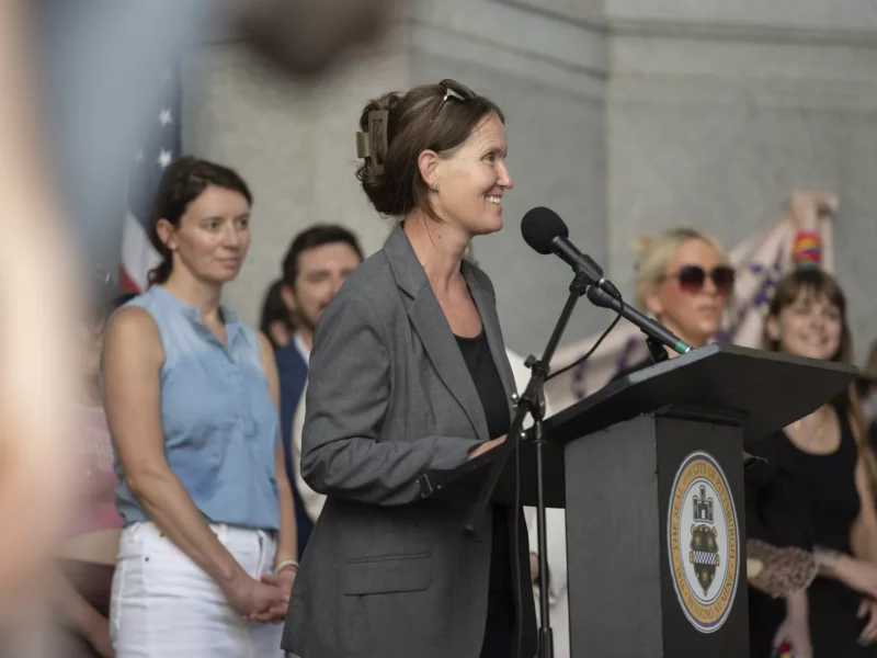 A woman in a business suit speaks under a high granite portico to a crowd of several dozen