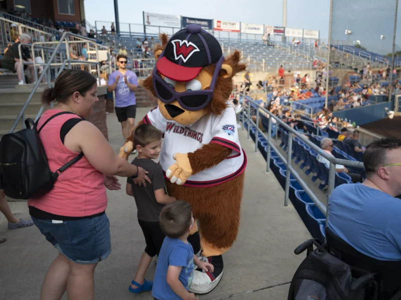 A large, wolflike mascot in a jersey and sunglasses hugs a young boy with encouragement from his mother