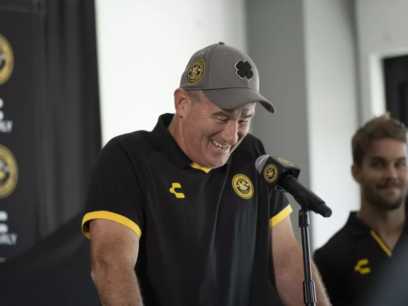 A clean-shaven man in a cap and soccer jersey smiles at a microphone and podium