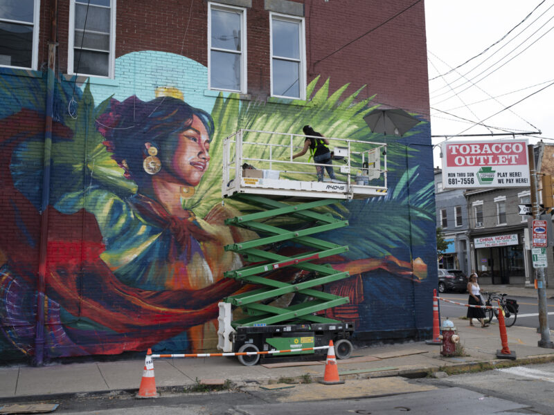 An artist works on a story-high mural of a woman dancing with lit candles