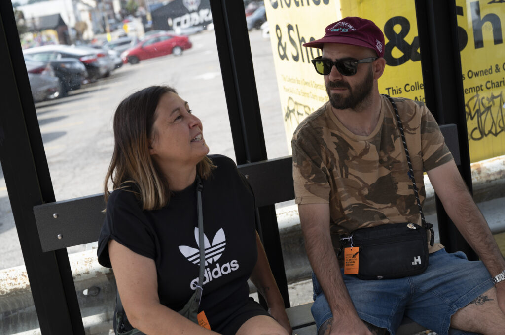 Two artists in casual clothing sit in a bus shelter