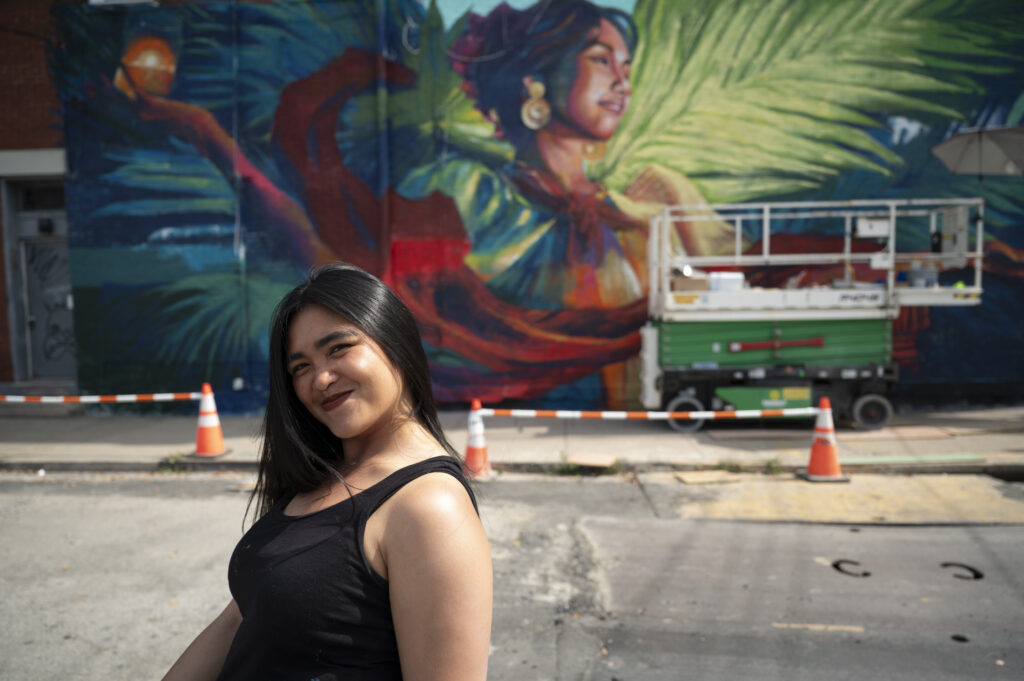 A woman with long straight hair smiles in a tanktop near a mural and scissor lift