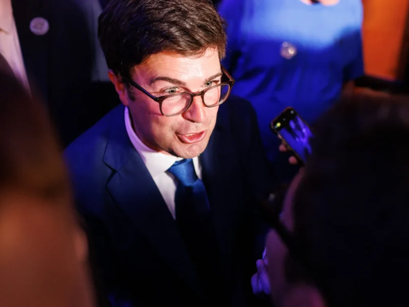 A clean-shaven man in a suit and glasses speaks animatedly to the media