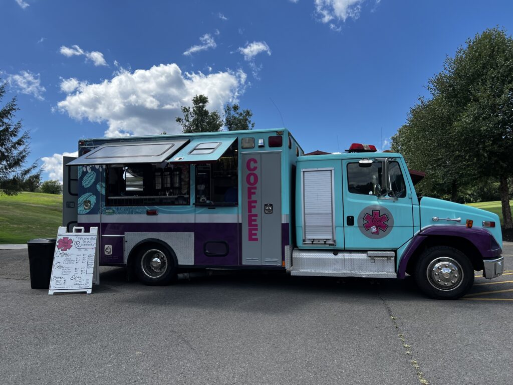 An ambulance with a window and awning on one side and a sandwich board announcing the day's specials