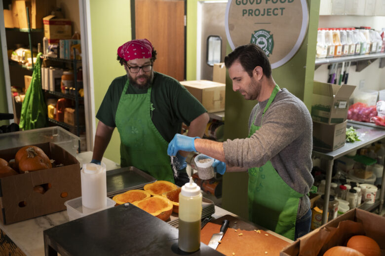 Two men in aprons stand over chopped pumpkin while the clean-shaven one sprinkles salt