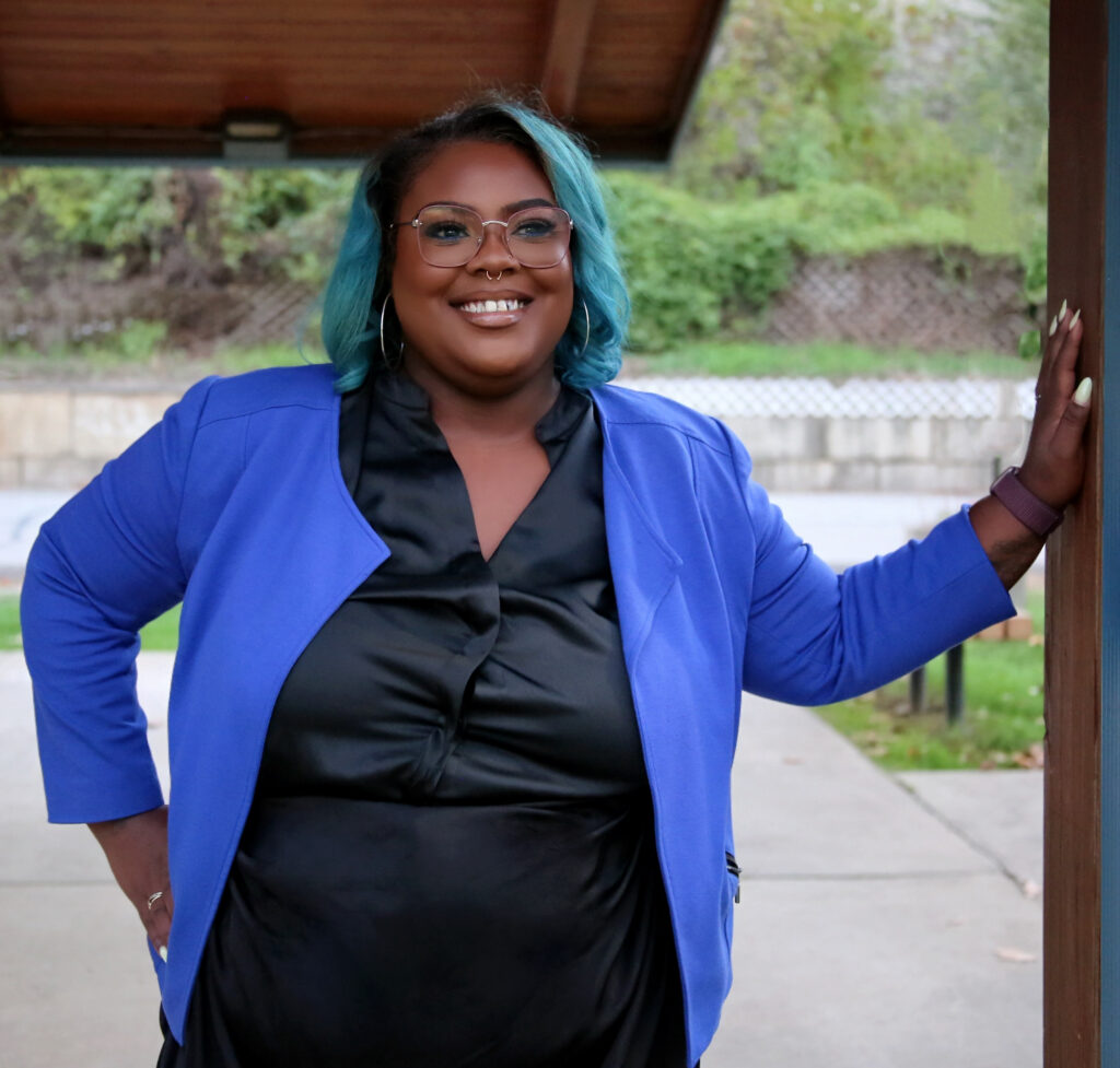 A black woman in a blouse and cardigan with blue hair and a septum ring leans against a pillar in an outdoor pavilion