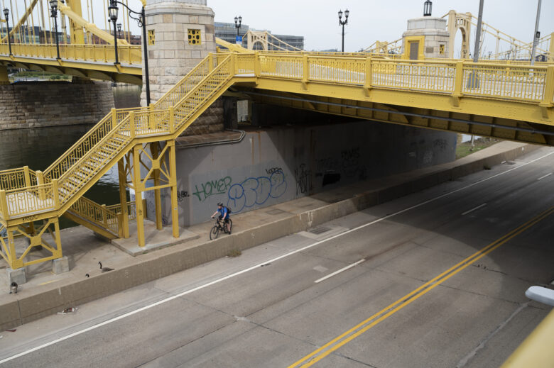 A narrow concrete bikeway and adjacent roadway pass beneath a suspension bridge and pedestrian stairs