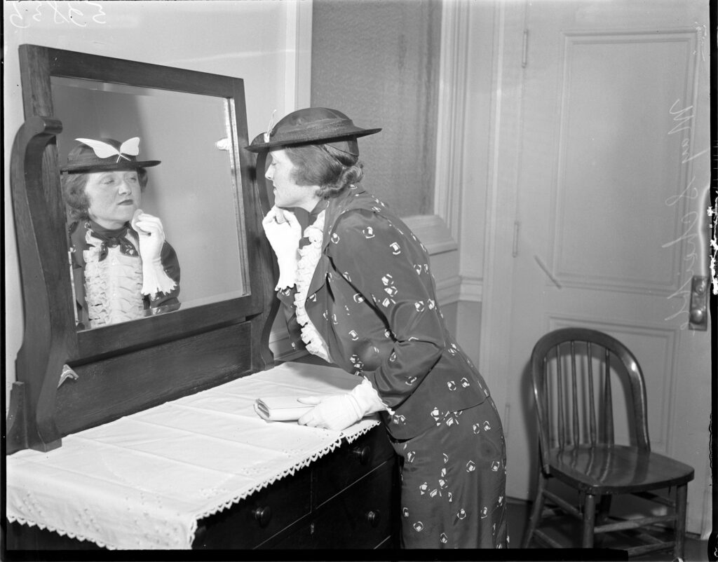 A woman in a hat and dress powders her face in a wooden mirror