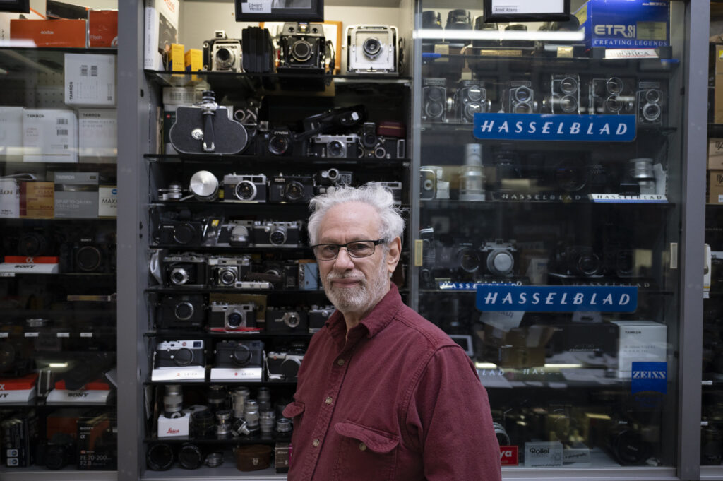 A man with glasses and a beard stands by a glass cabinet full of vintage Hasselblad cameras