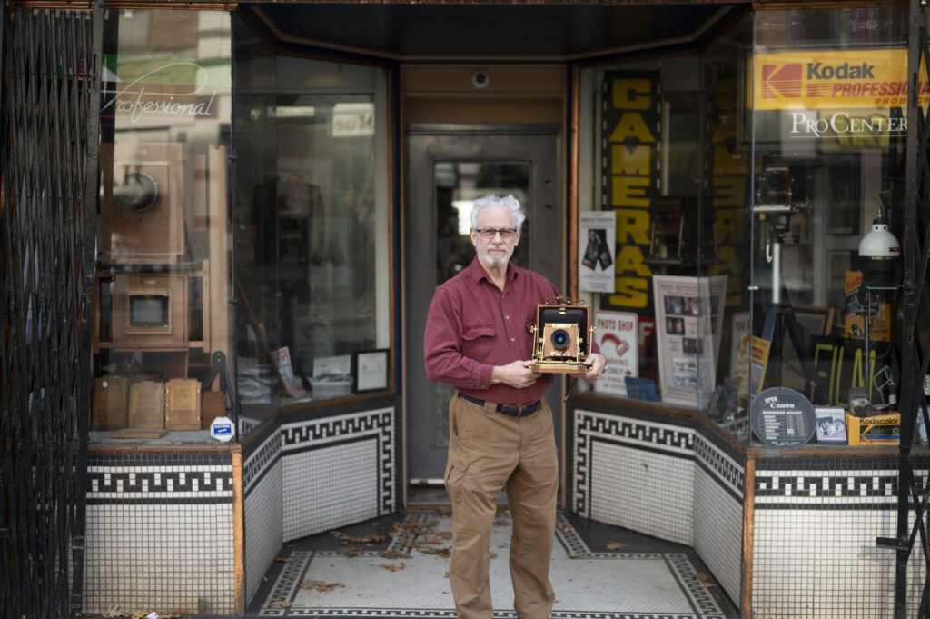 A man with glasses and a beard holds an antique camera outside a Victorian storefront