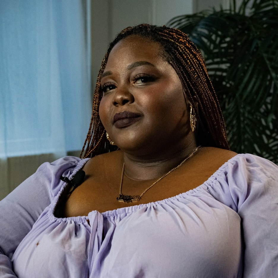 A woman with long braids wearing a loose lavender blouse, necklace, and earrings